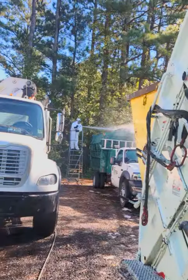 Crew member in protective suit washing a garbage truck on site in a wooded yard