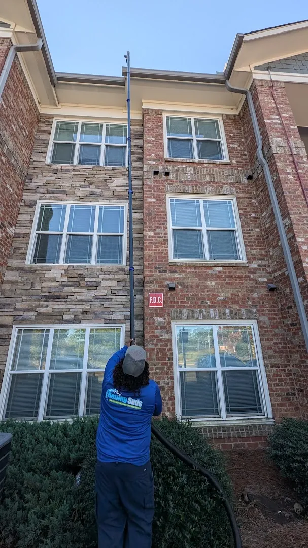 Rolling Suds technician vacuuming third-story gutters on a brick apartment community building from the ground